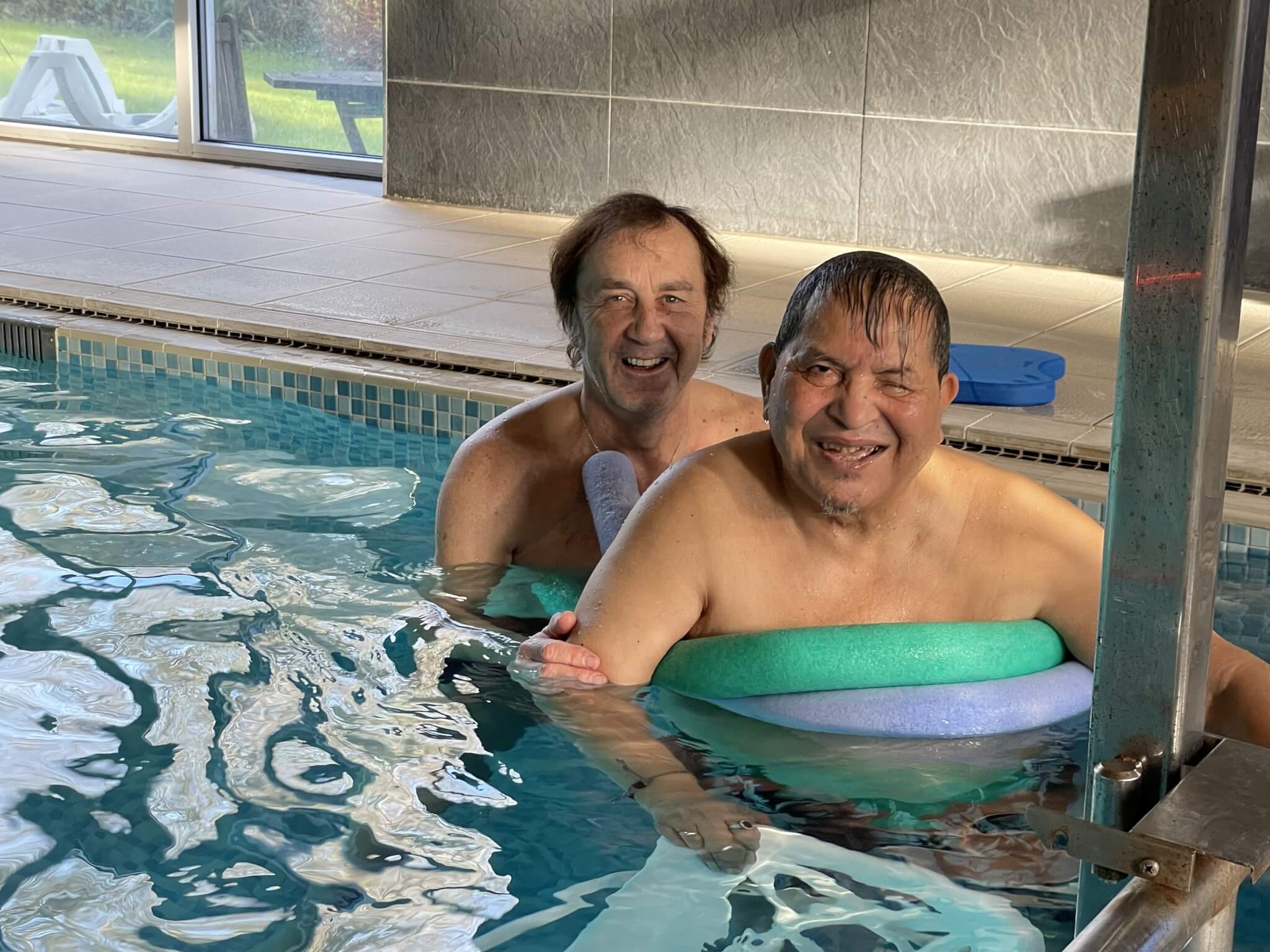 Two smiling men in a swimming pool, one wearing a blue and green pool noodle, with windows and tiled walls in the background. - Home Instead