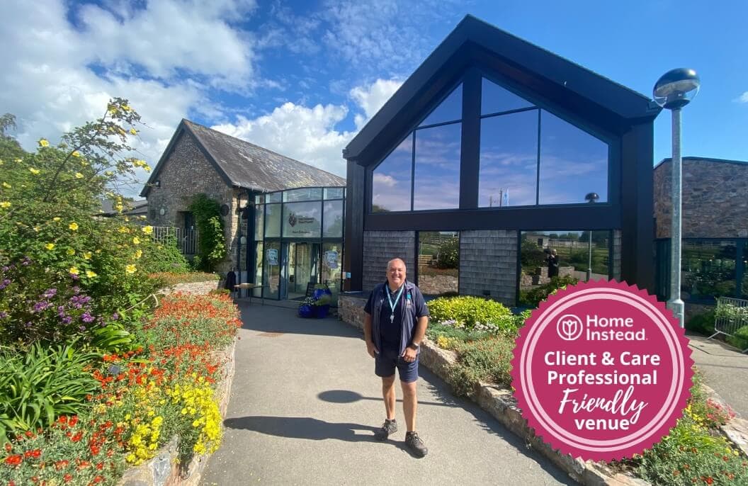 A person stands outside a modern building with a sign reading "Home Instead - Client & Care Professional Friendly Venue. - Home Instead