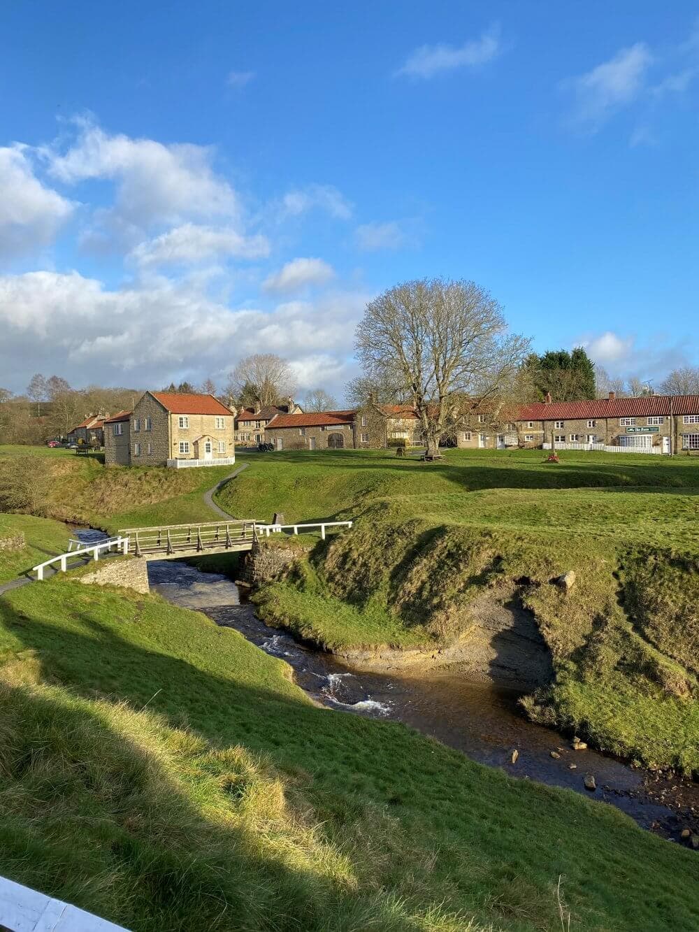 Scenic countryside with a stream, small bridge, and houses under a clear blue sky with a few clouds. - Home Instead