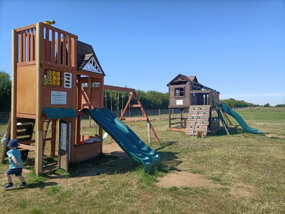 Child playing near wooden playground structures with slides and swings in a spacious grassy field under a clear blue sky. - Home Instead
