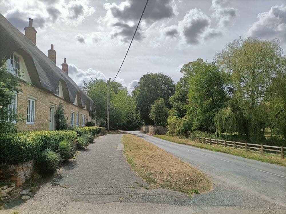 A rural road beside charming thatched cottages and lush greenery under a partly cloudy sky. - Home Instead