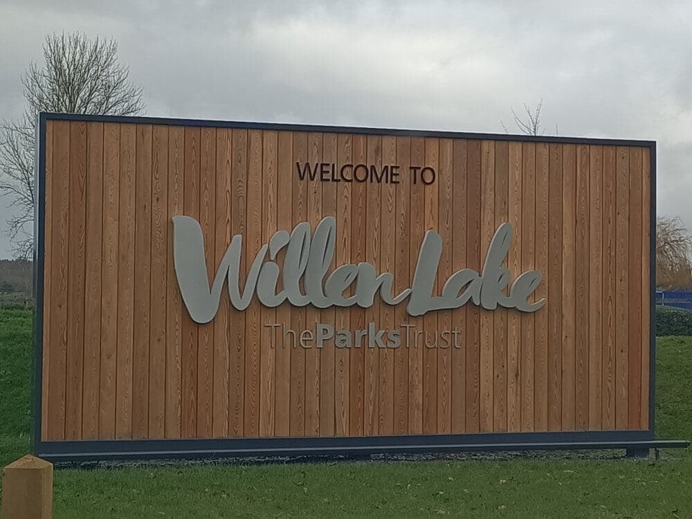 A wooden sign that reads "Welcome to Willen Lake, The Parks Trust" on a grassy field with a cloudy sky. - Home Instead