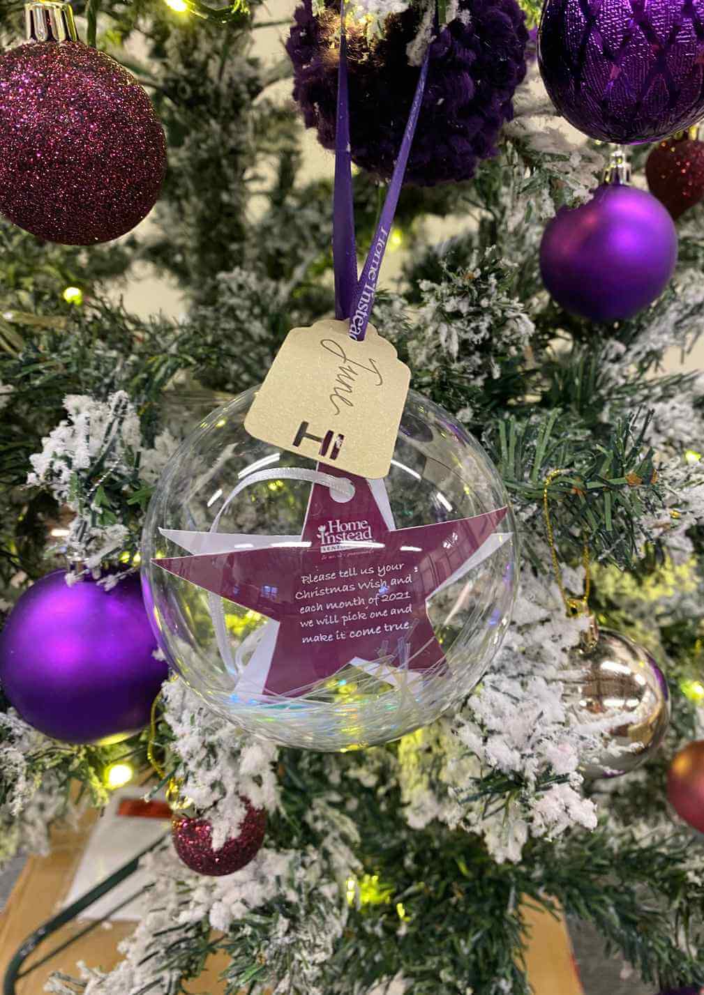 A Christmas tree adorned with purple ornaments and a transparent ornament with a wish tag hanging from it. - Home Instead