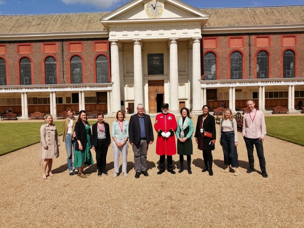 Group of people, including a person in a red uniform, standing outside a large building with columns and arched windows. - Home Instead