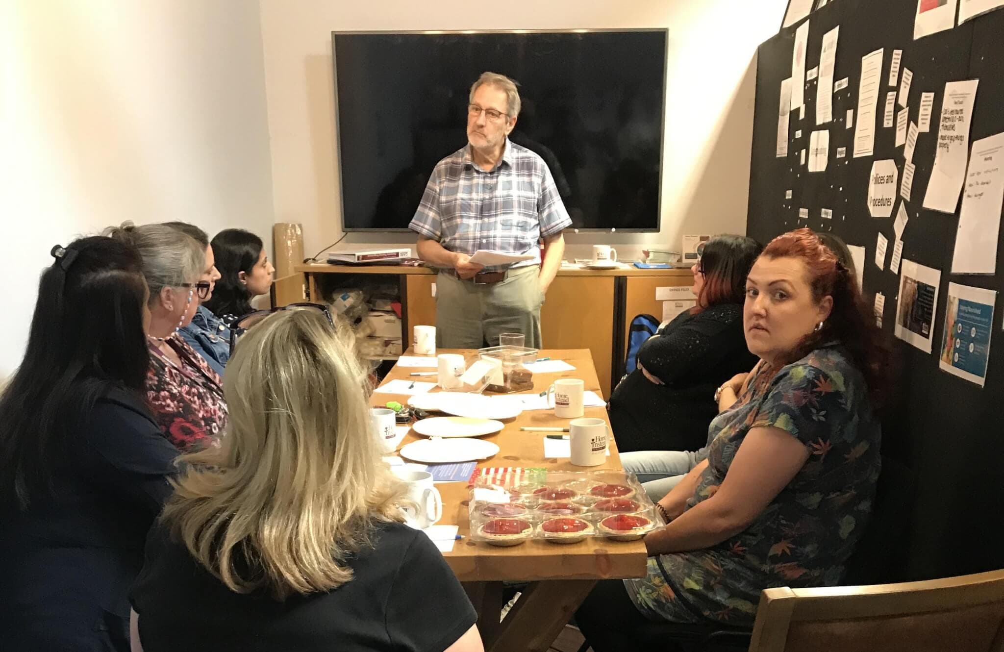A man stands in front of a group of people seated at a table, giving a presentation in a meeting room. - Home Instead