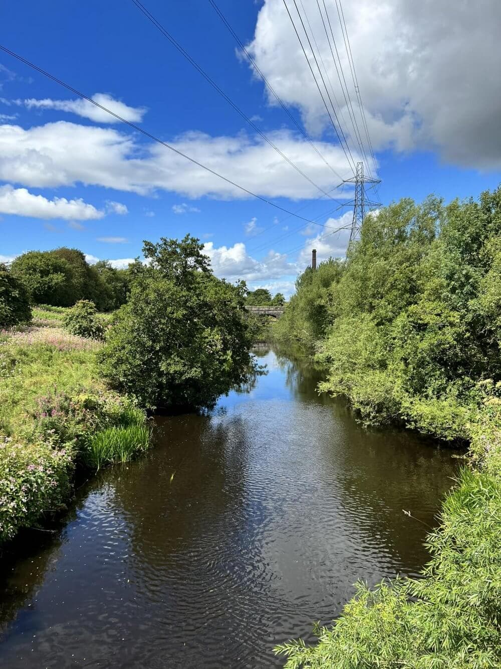 Jenny's Well - A serene river flows through lush greenery with power lines overhead and a partly cloudy blue sky above. - Home Instead Renfrewshire and Barrhead home care services Paisley