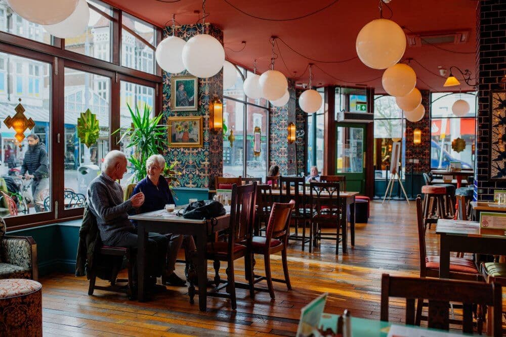 Two elderly people sitting at a table in a cozy cafe with large windows and modern decor. - Home Instead