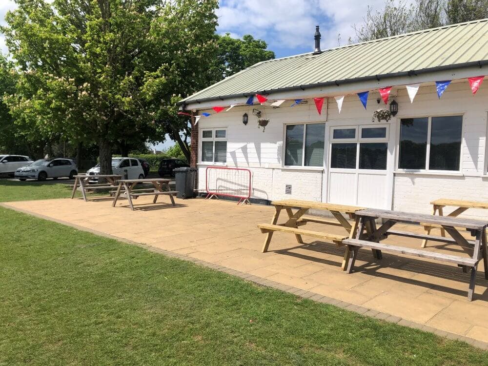 A small building with a green roof, picnic tables, and colorful bunting, bordered by trees and parked cars. - Home Instead