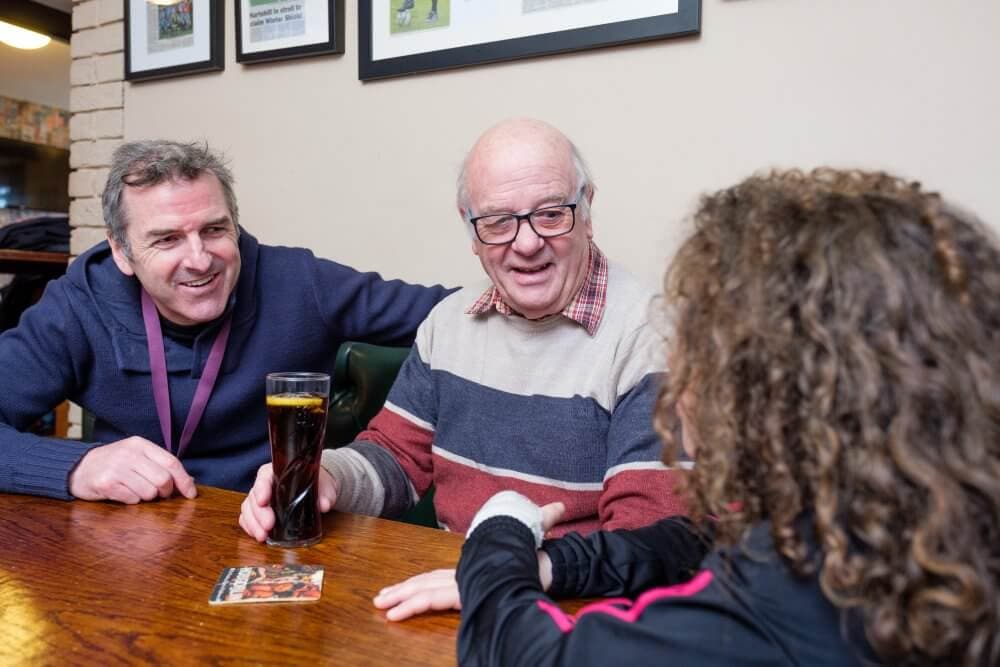 Three people smiling and chatting at a table, one holding a drink, in a cozy setting with framed pictures on the wall. - Home Instead
