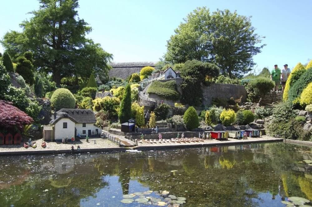 A lush, scenic village with miniature houses beside a pond reflecting the greenery and people exploring the area. - Home Instead