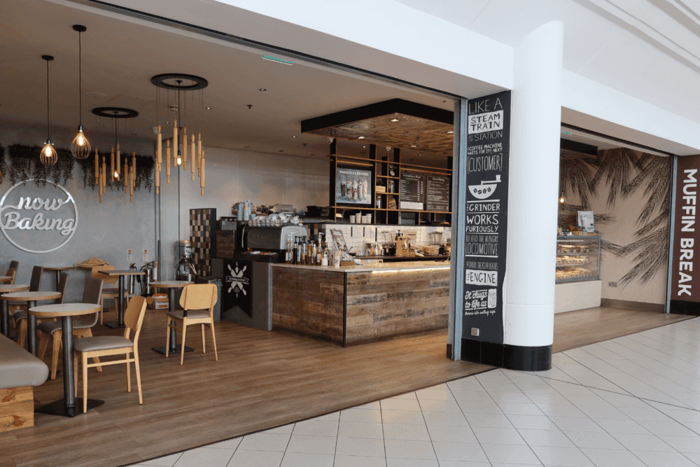 A cozy café with wooden decor, a "Now Baking" neon sign, hanging lights, and baked goods on display. - Home Instead