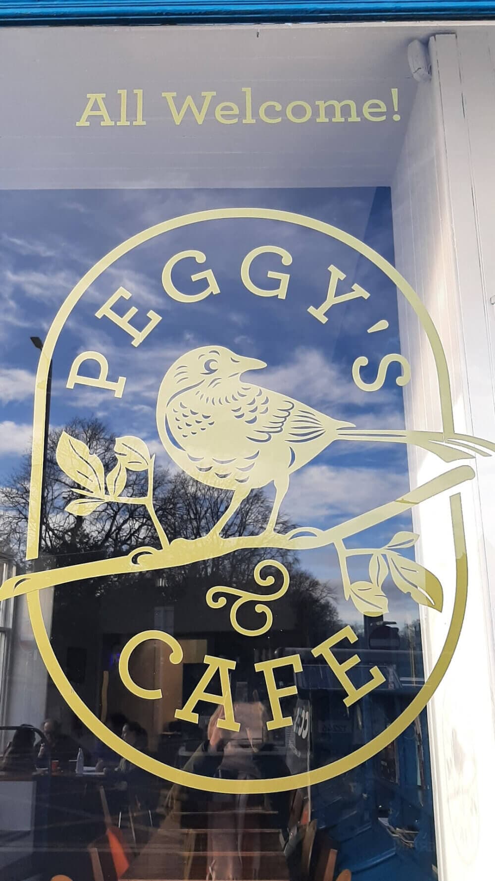 A window sign featuring a bird on a branch and the text "PEGGY'S CAFE" with "All Welcome!" above. - Home Instead