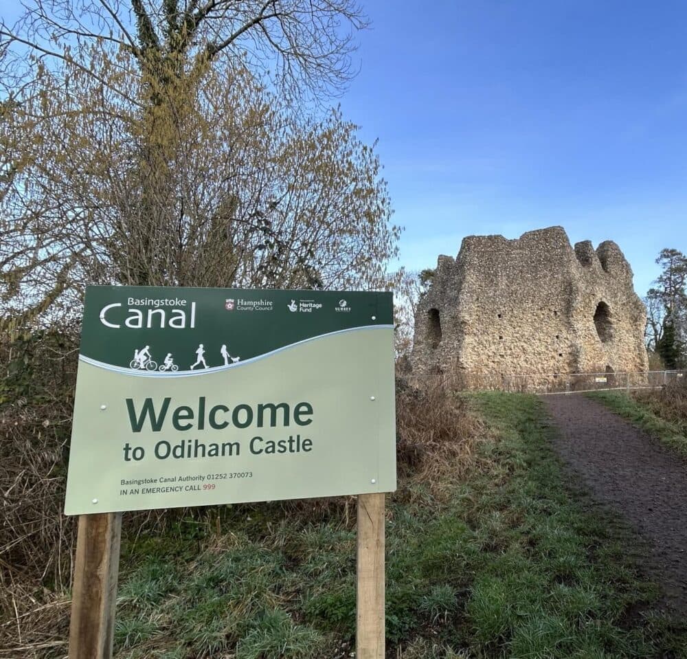 Picture of a sign in front of a pathway leading to the ruins of Odiham Castle, surrounded by trees and greenery.