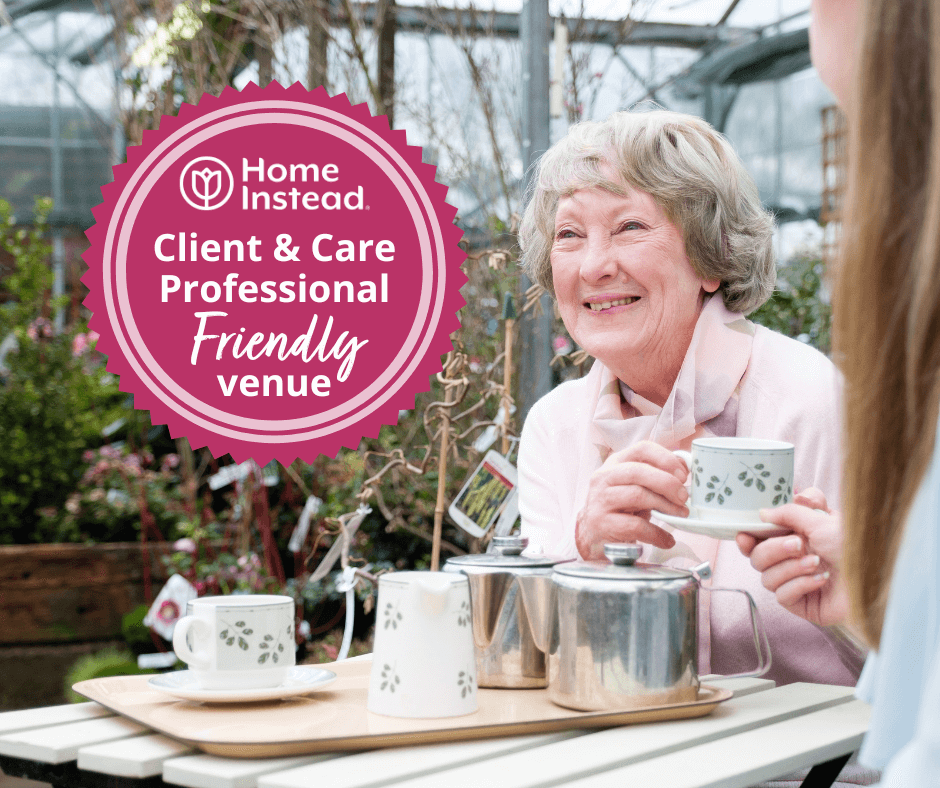 Elderly woman smiles while drinking tea with another person, badge reads "Home Instead, Client & Care Professional Friendly Venue. - Home Instead