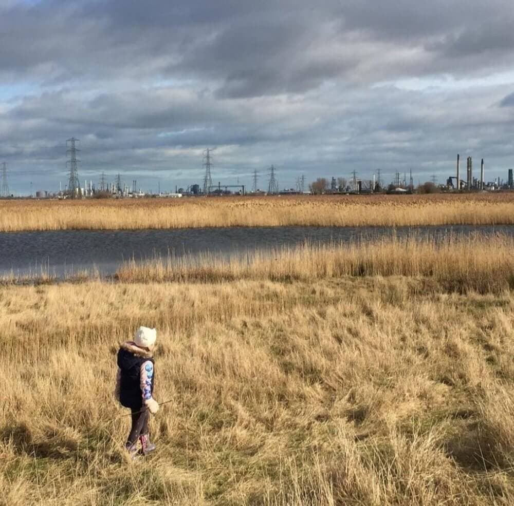 A child walks through a grassy field with a river and industrial structures under a cloudy sky in the background. - Home Instead