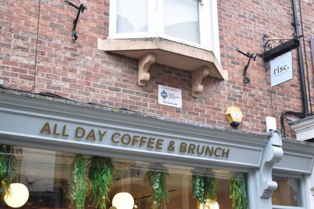 A café with a sign "ALL DAY COFFEE & BRUNCH," plant decor, and brick exterior under a window on an upper floor. - Home Instead