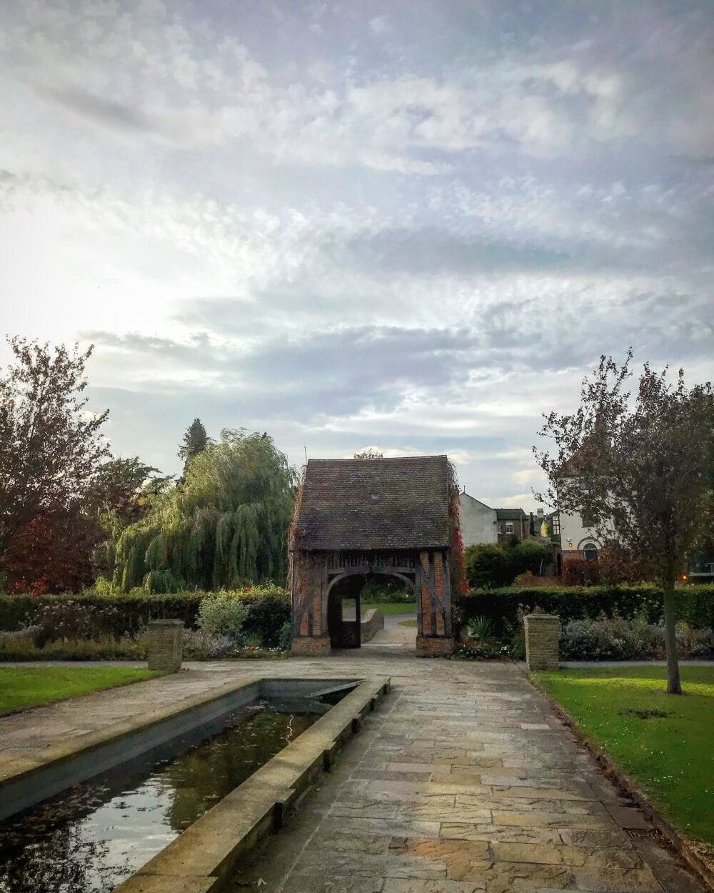 A charming archway entrance in a lush garden with a reflecting pool and cloudy sky. - Home Instead