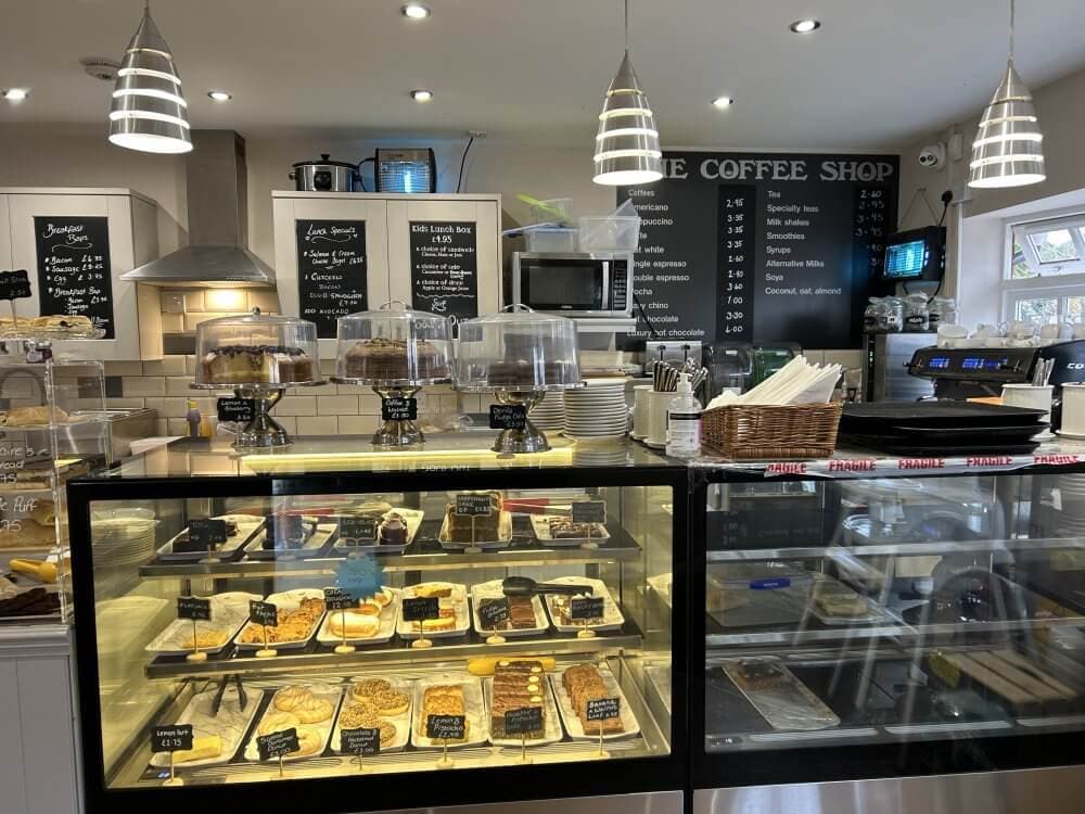 Home Instead Basingstoke picture of The Coffee Shop at Sherfield on Loddon, showing a glass display of various pastries, cakes, and sandwiches with chalk menu boards and hanging lights above the counter.
