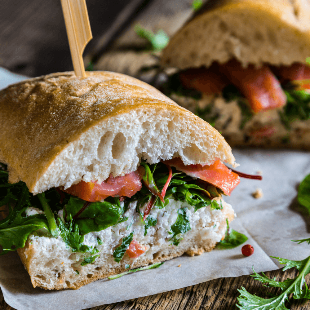 Close-up of a fresh sandwich filled with greens, tomatoes, and ricotta cheese on parchment paper with a wooden skewer. - Home Instead