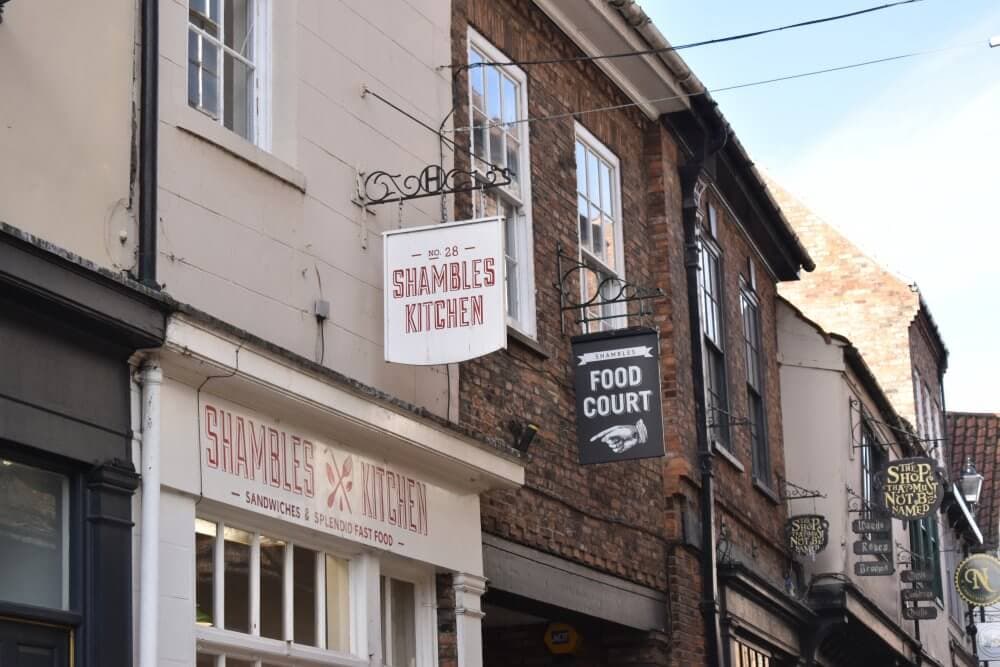 A street view shows signs for "SHAMBLES KITCHEN," "FOOD COURT," and nearby shops against a clear sky. - Home Instead