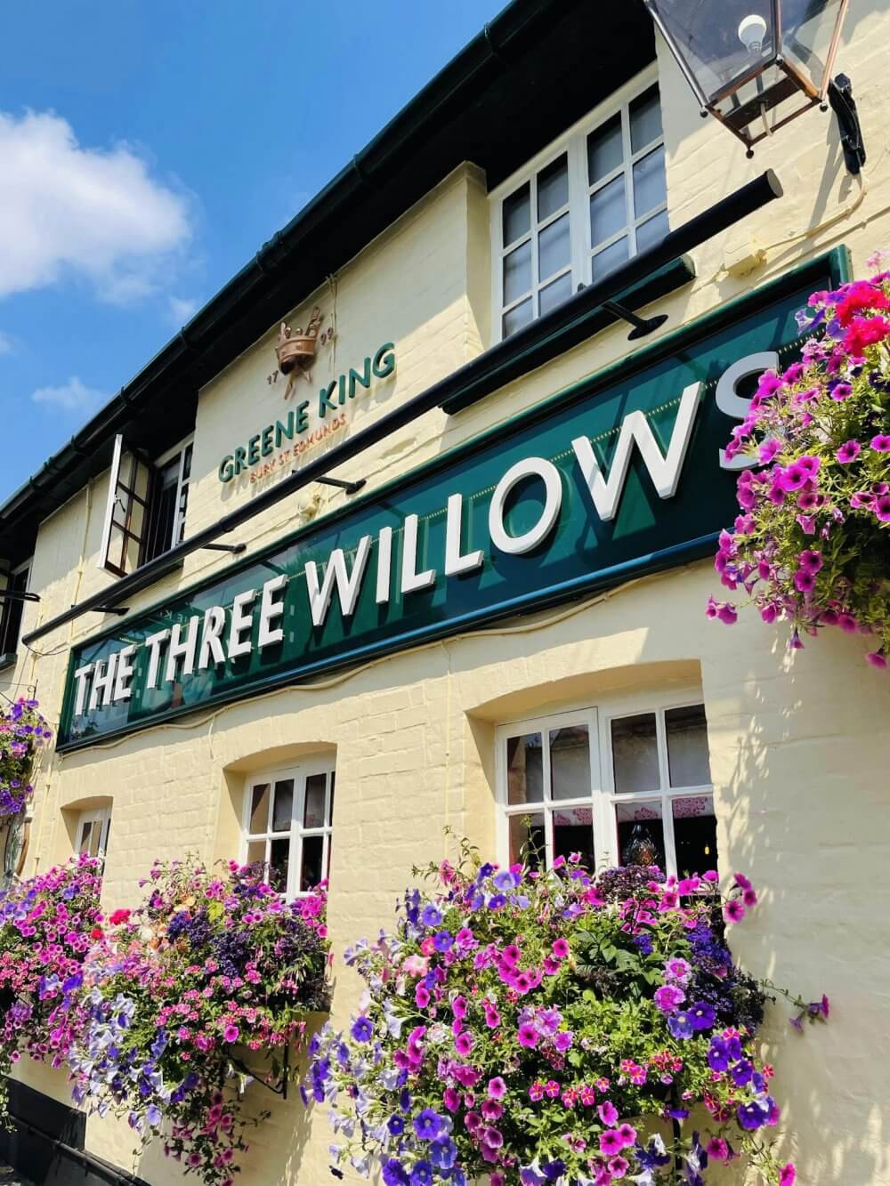 Exterior of a pub called "The Three Willows" with hanging flower baskets and signage reading "Greene King. - Home Instead