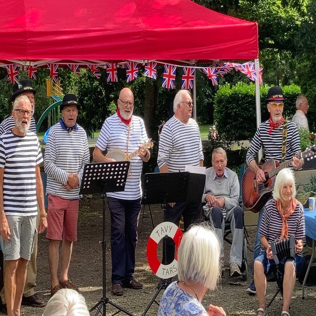 A group of older adults in striped shirts perform music under a red tent decorated with Union Jack bunting. - Home Instead