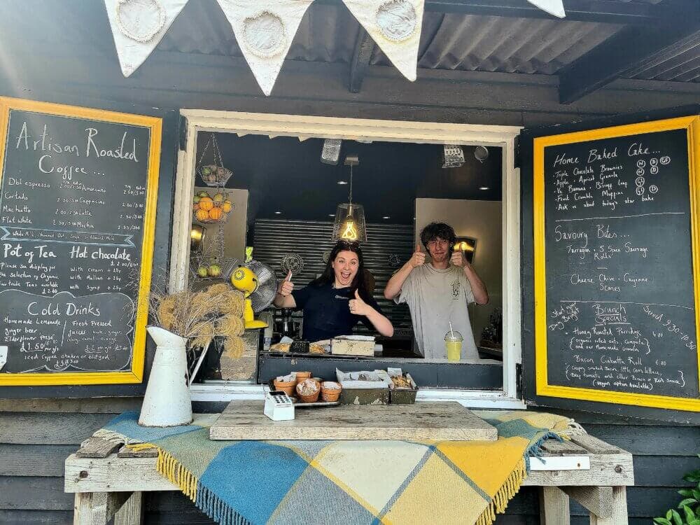 Home Instead Basingstoke picture of The Hobo café at Hortus Loci garden centre, near Hook, showing two people smiling and giving thumbs up inside a café, with chalkboard menus and various items on the counter.