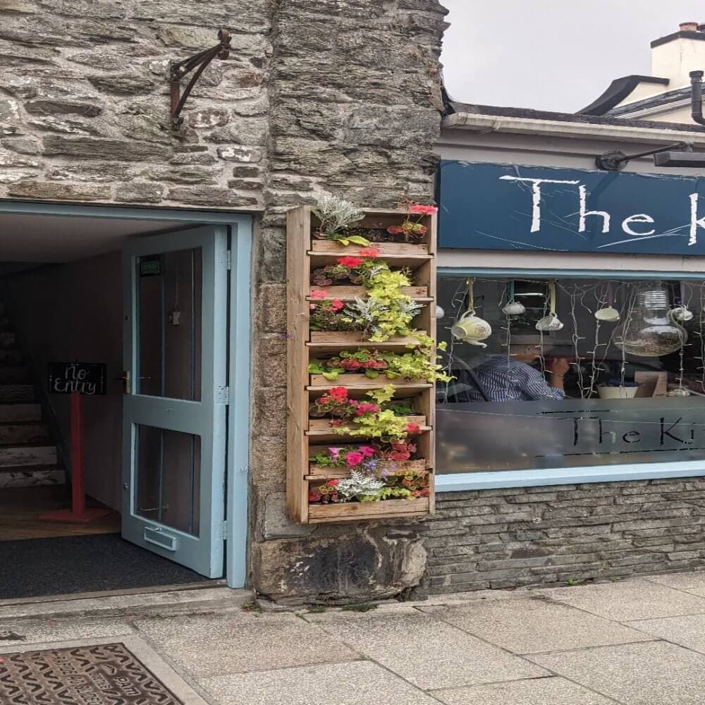 Stone building with blue door, vertical flower garden, and window displaying hanging ornaments with "The" sign. - Home Instead