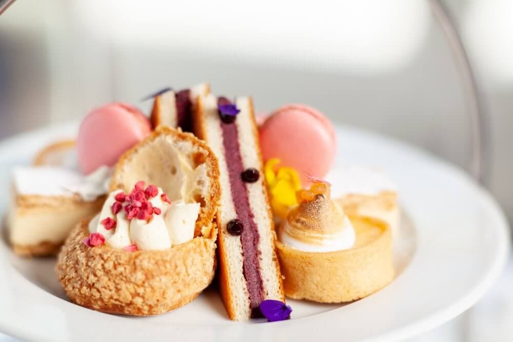 A plate of assorted pastries including macarons, cake slices, and tarts, garnished with cream and flowers. - Home Instead