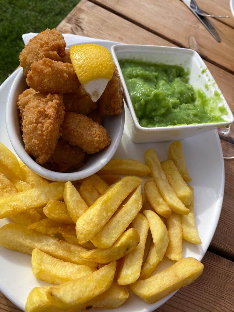 A plate of fries, crispy chicken bites with a lemon wedge, and a side of mushy peas served outdoors. - Home Instead