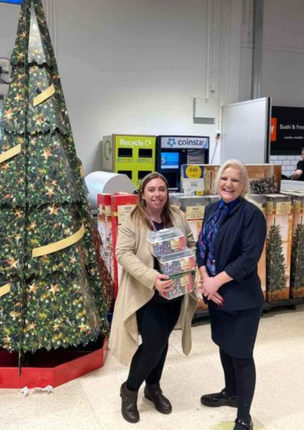 Two women stand by a decorated Christmas tree, holding festive tins, in a store with a Coinstar machine visible. - Home Instead