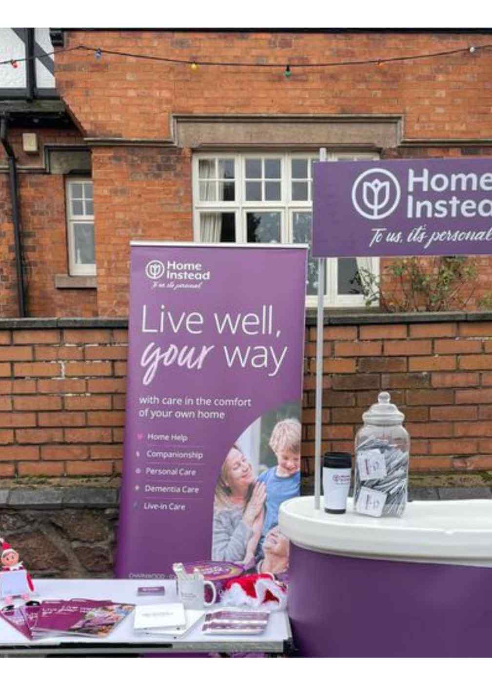 Outdoor table setup with banners for Home Instead, offering home help, companionship, personal care, and live-in care services. - Home Instead