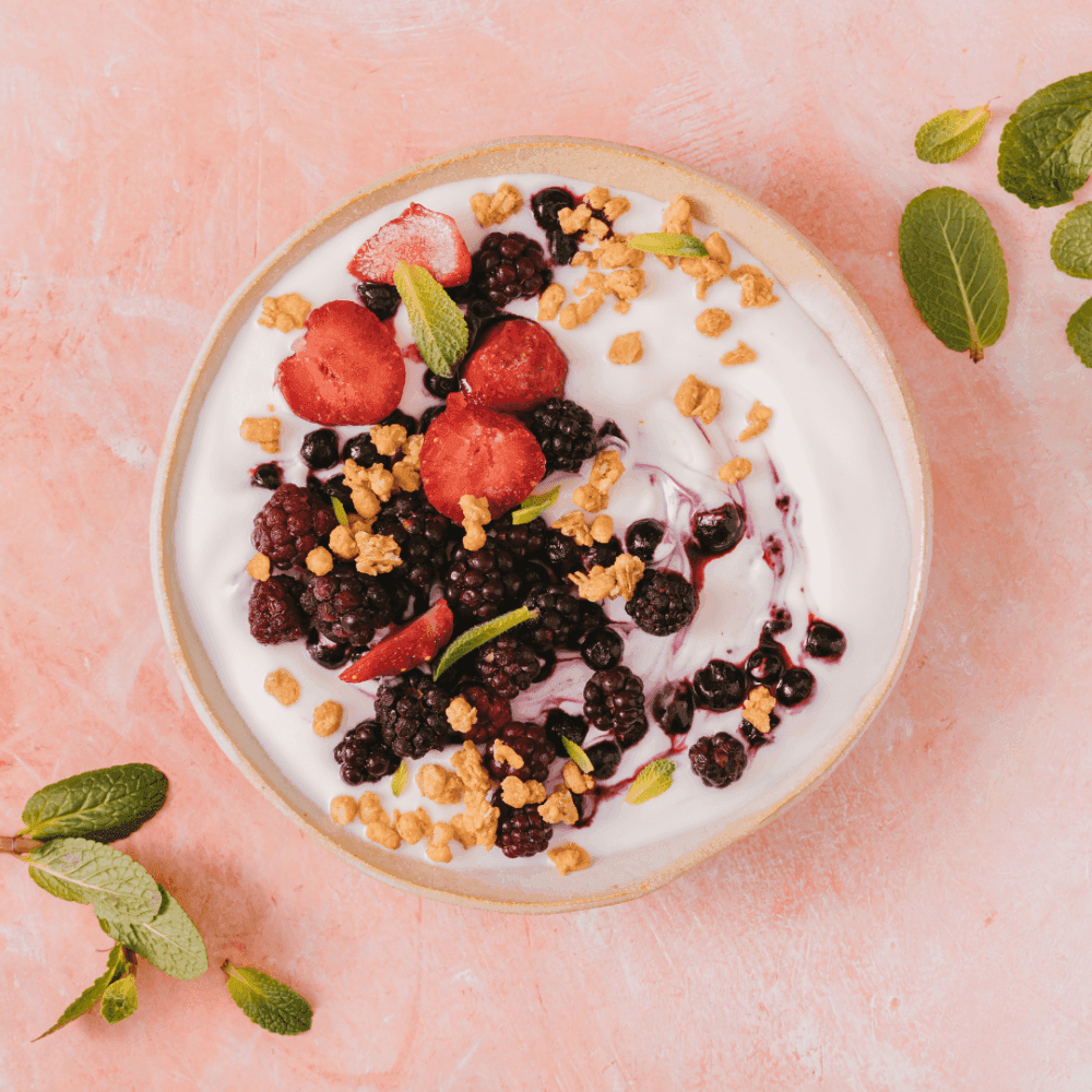 Bowl of yogurt topped with strawberries, blackberries, granola, and mint leaves on a pink surface, with mint nearby. - Home Instead