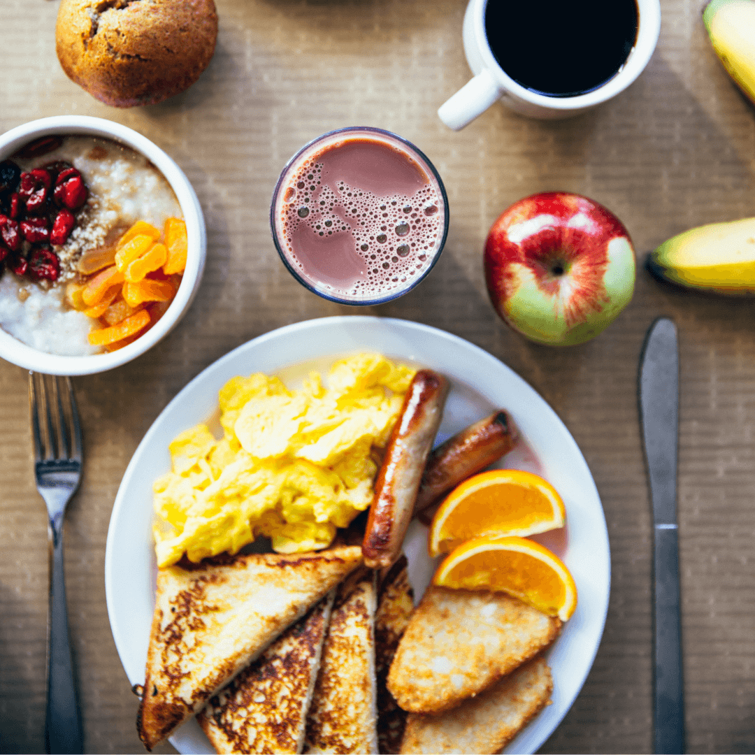 A breakfast spread with scrambled eggs, toast, sausages, orange slices, oatmeal, a muffin, coffee, chocolate milk, and fruit. - Home Instead