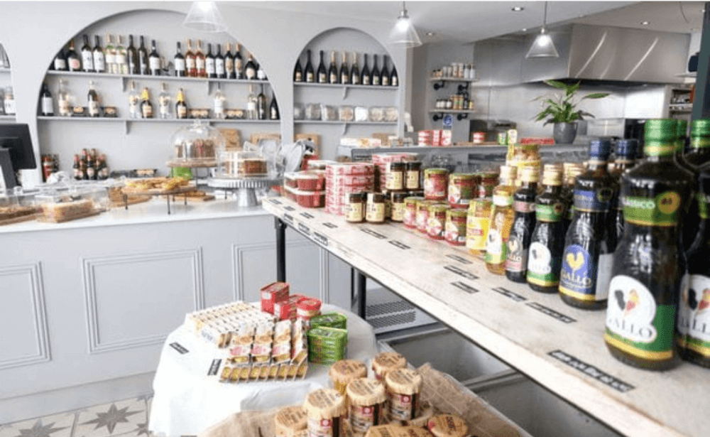 Interior of a gourmet grocery store with shelves displaying various wines, preserves, oils, and breads in a bright setting. - Home Instead