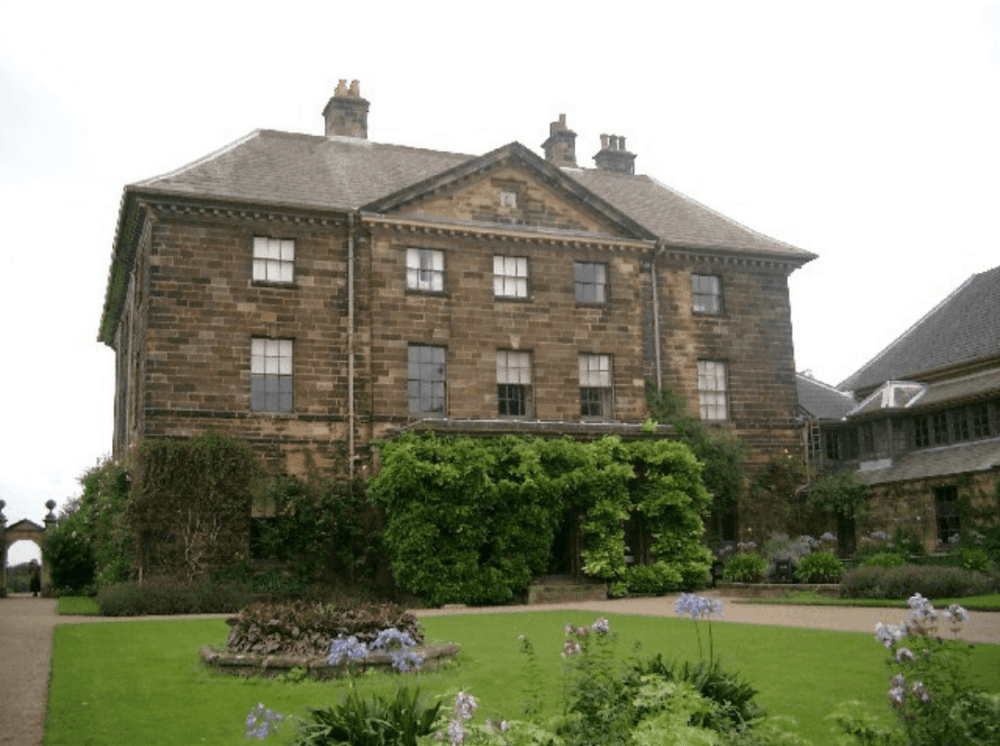 A large, historic, two-story stone building with lush green foliage and a manicured garden in the foreground. - Home Instead