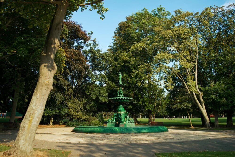 A green fountain surrounded by trees in a sunny park. There is a dirt path leading to the fountain. - Home Instead