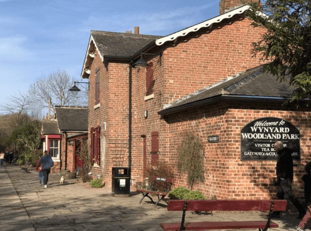A person walks a dog next to a brick building with a sign reading "Welcome to Wynyard Woodland Park. - Home Instead