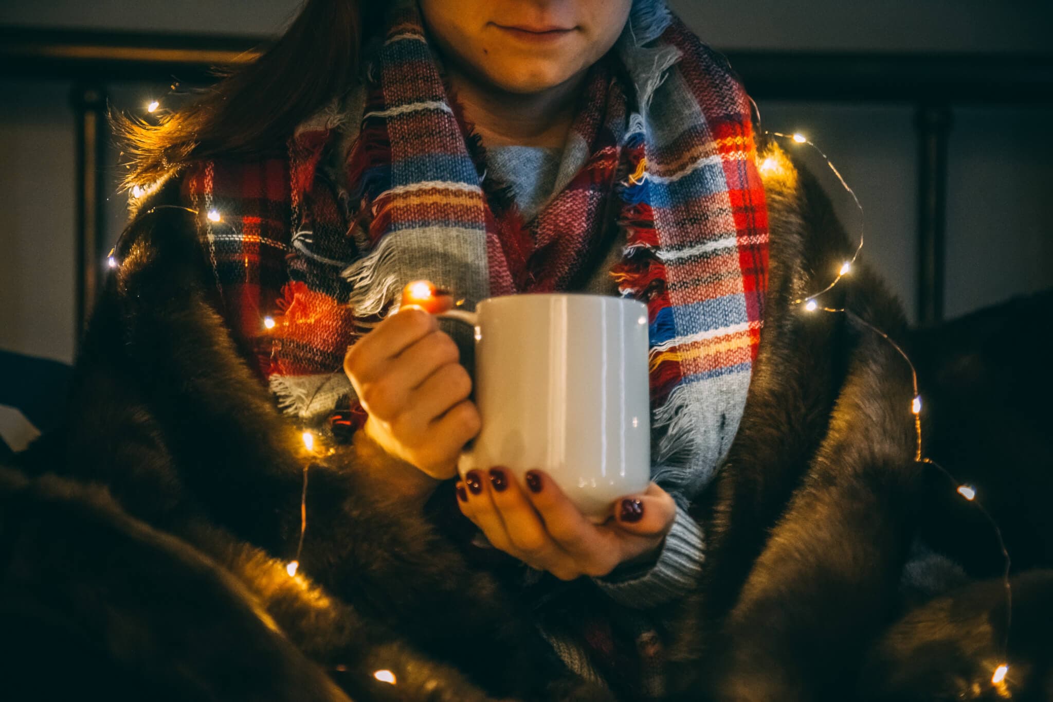 Person holding a white mug, wrapped in a plaid blanket and fairy lights, cozy winter scene. - Home Instead