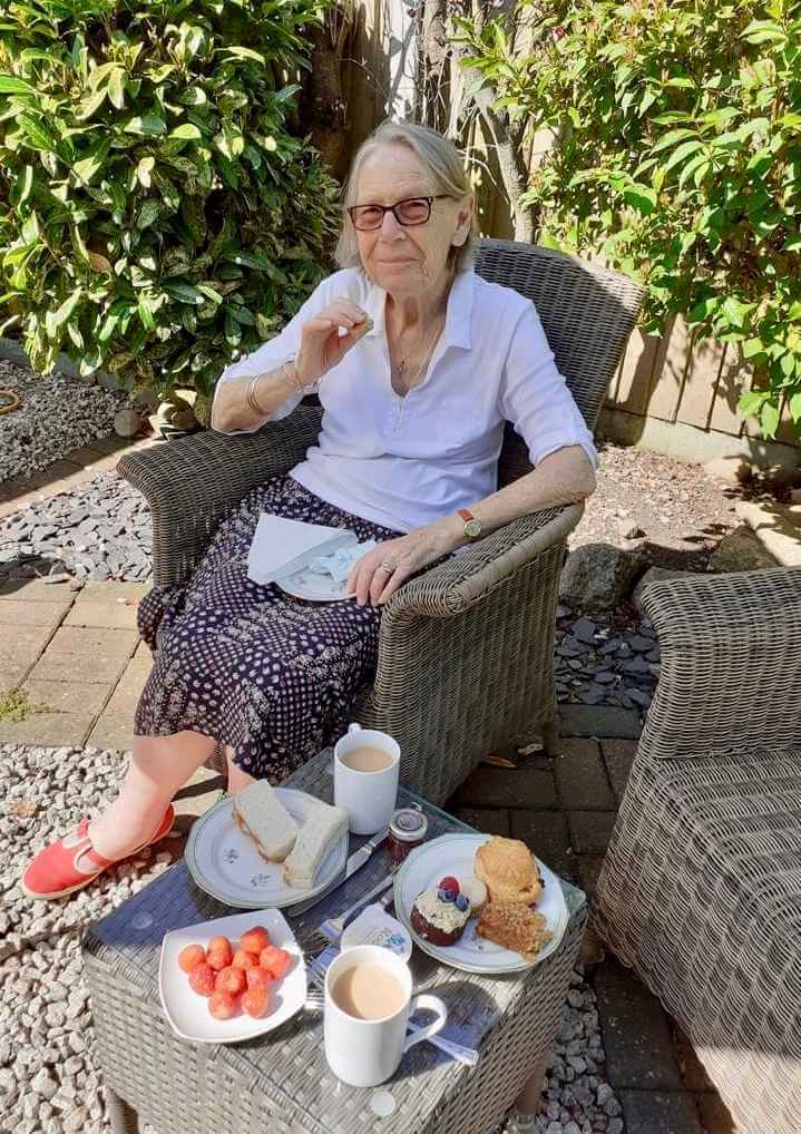 An elderly woman sits outdoors at a table with tea, sandwiches, scones, and strawberries, smiling in a sunny garden. - Home Instead