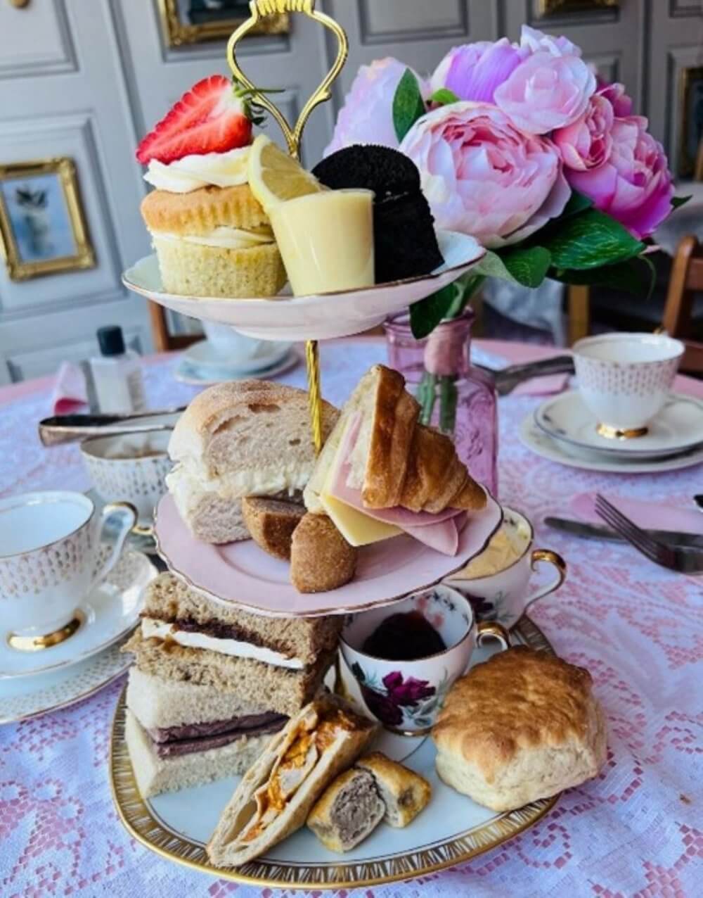 A tiered tray with assorted tea sandwiches, pastries, and desserts, beside a teapot and teacups, near a vase with flowers. - Home Instead