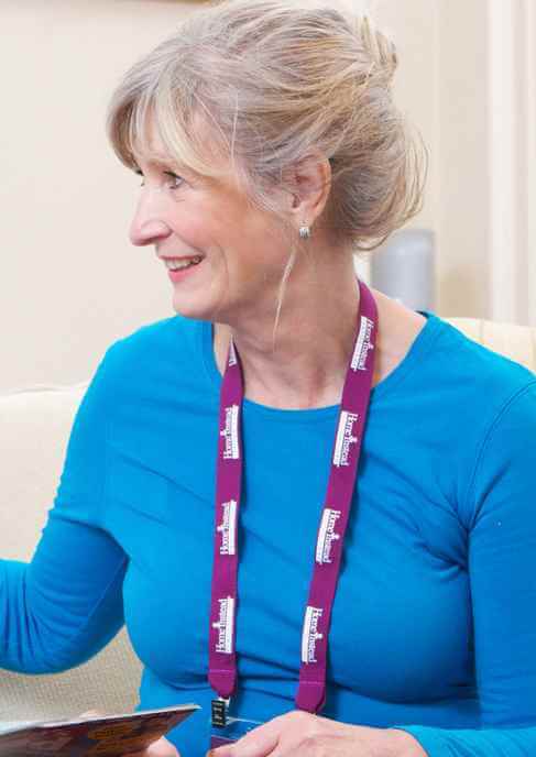 Person with gray hair in a bun, wearing a blue top and a purple lanyard, smiling and looking to the side. - Home Instead