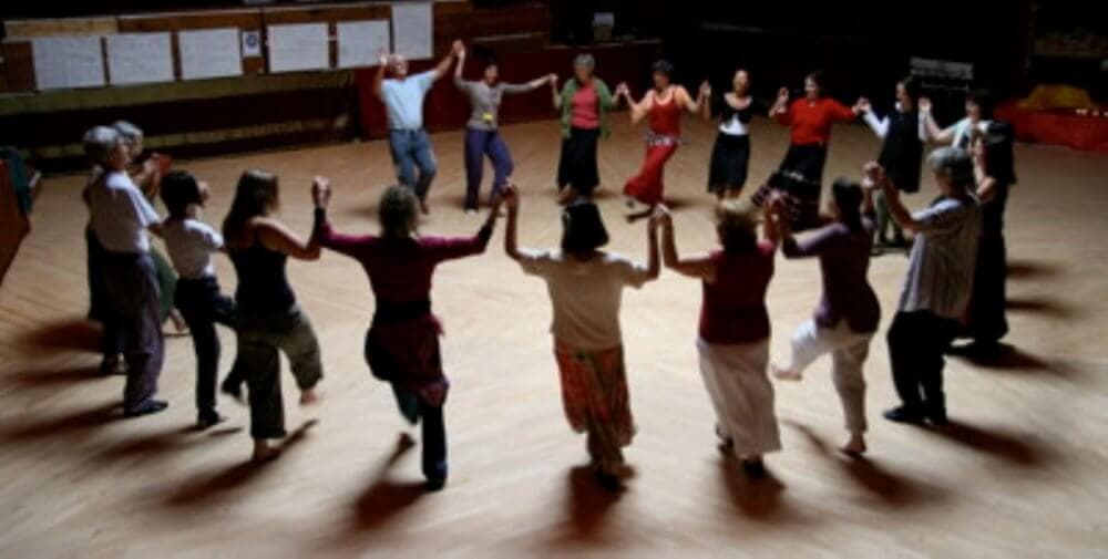 A group of people holding hands and dancing in a circle indoors. - Home Instead