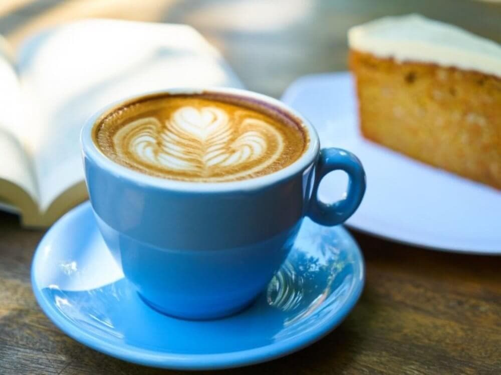 A cup of latte with latte art in a blue cup and saucer, next to an open book and a slice of cake on a plate. - Home Instead
