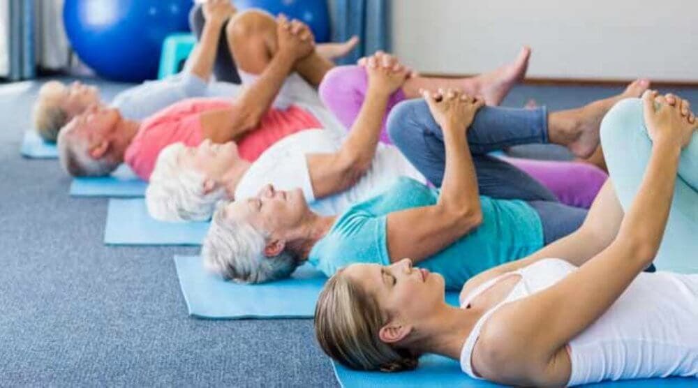 People lying on yoga mats, stretching their legs to their chest, in a group exercise session. - Home Instead