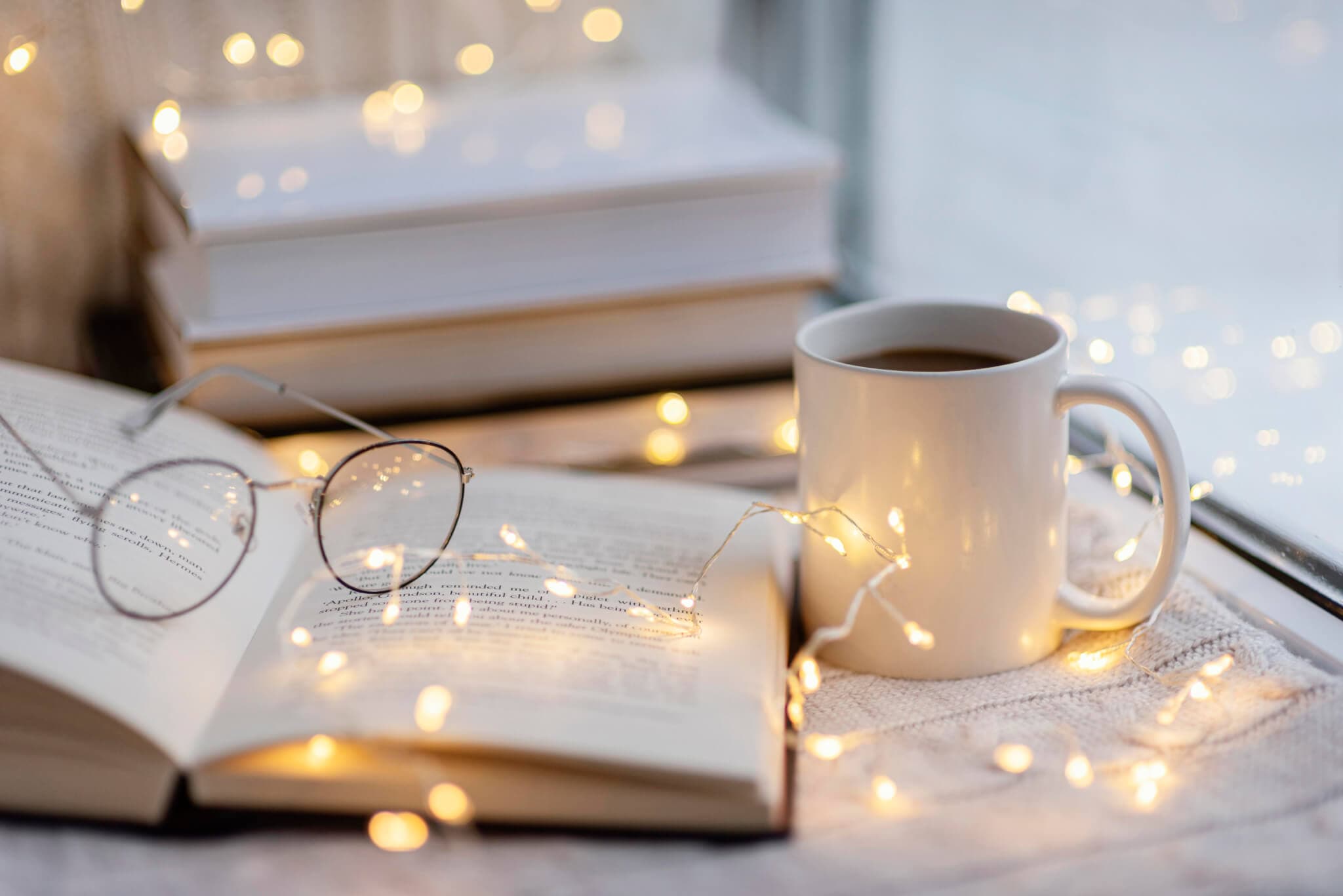 Open book with glasses, a mug of coffee, and string lights creating a cozy ambiance by the window. - Home Instead Renfrewshire and Barrhead Care at Home Services