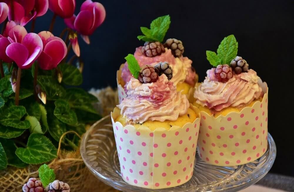 Three decorated cupcakes with pastel frosting and berries on a glass stand, next to a blooming pink cyclamen plant. - Home Instead