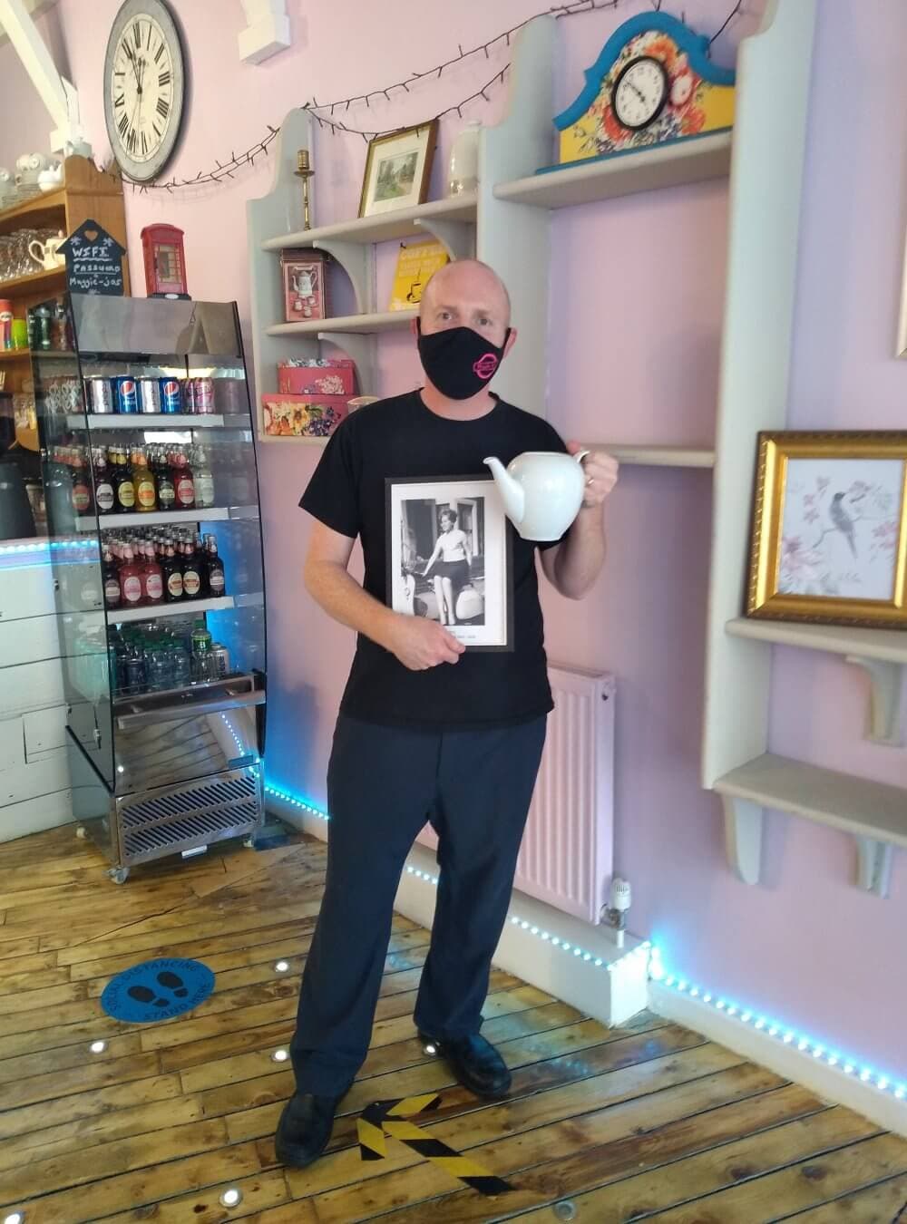 A man wearing a mask holds a framed photo and a large white teapot inside a decorated room with shelves and drinks. - Home Instead