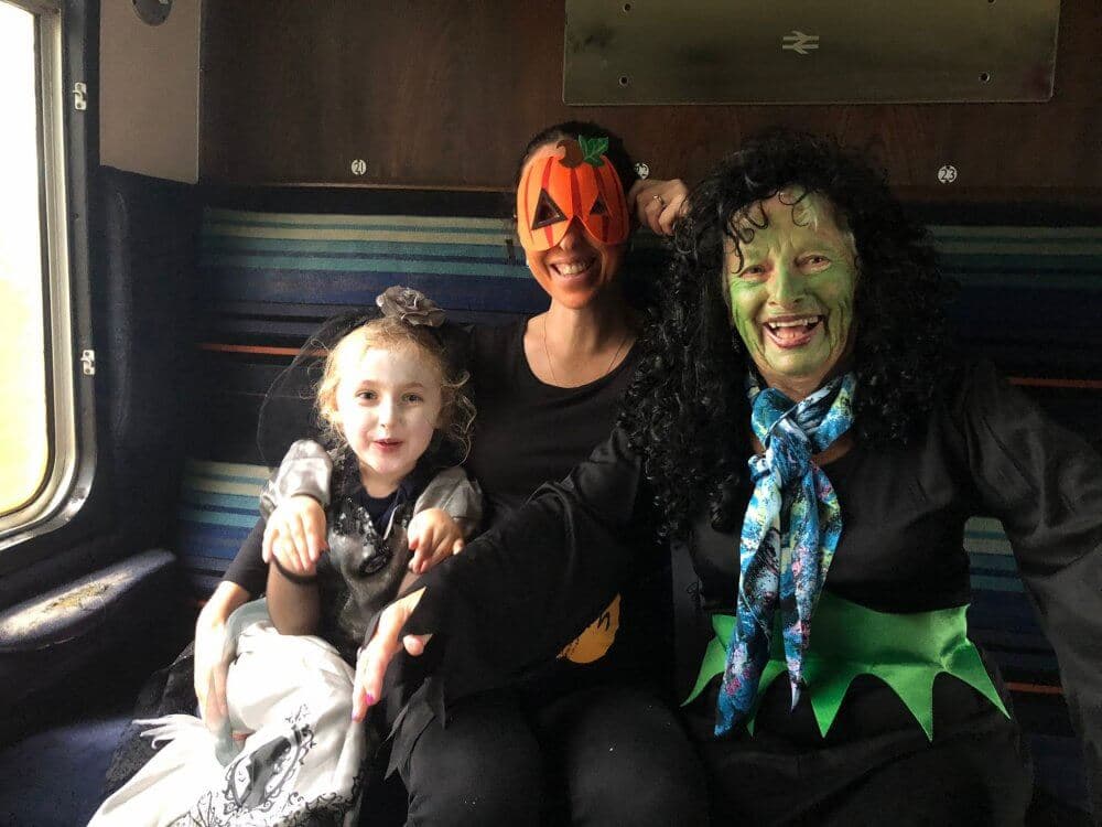 Three people in Halloween costumes sitting on a train; child as a princess, adult as a pumpkin, and adult as a green-faced witch. - Home Instead
