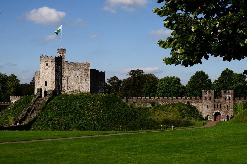 Cardiff Castle with a flag atop, lush green grounds, blue sky, and trees in the background. - Home Instead
